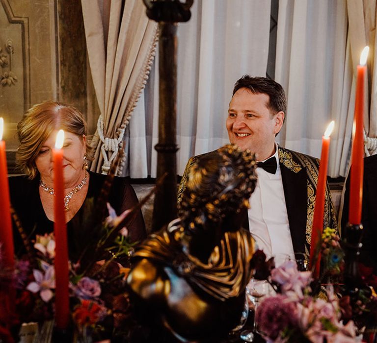 Groom in black and gold suit jacket with groom in black tuxedo smiling as they listen to wedding speeches 