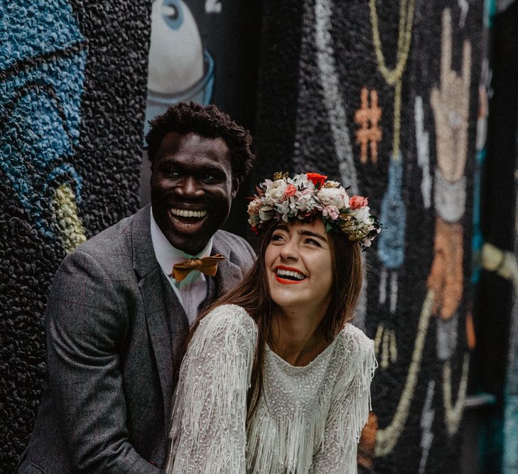 White bride wearing floral crown and Black groom wearing tweed suit laugh together against graffiti wall