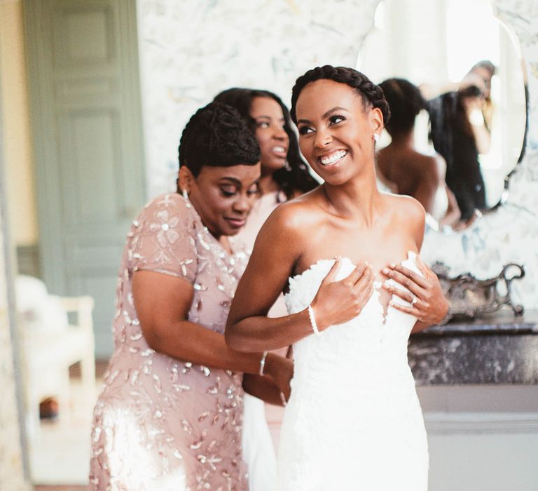 Bridesmaids wearing pink help to dress a bride on the morning of the wedding. The bride holds her dress close whilst laughing.