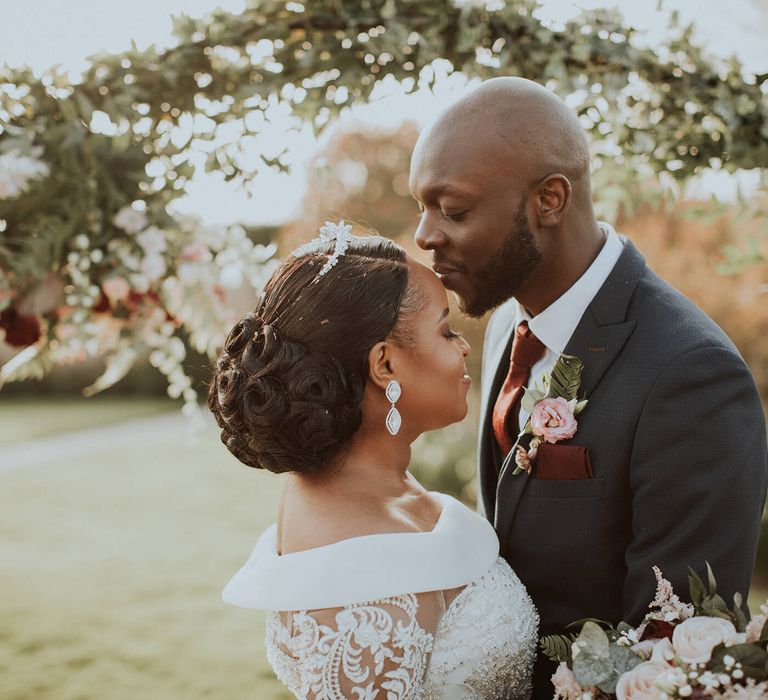 A groom kisses his bride on the forehead. The bride has a low bun in her hair and and off the shoulder dress.