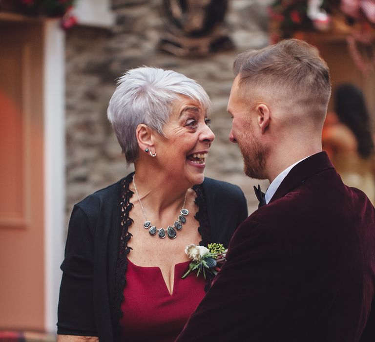 Groom in a burgundy velvet jacket talking to a wedding guest