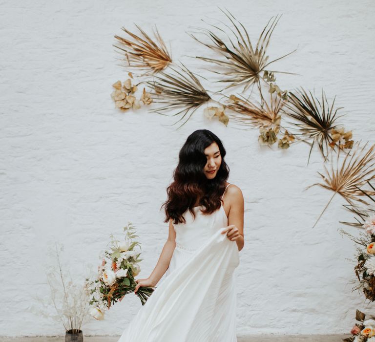 The bride swishing around the pleated skirt of her wedding dress in front of an arch of dried palm leaves