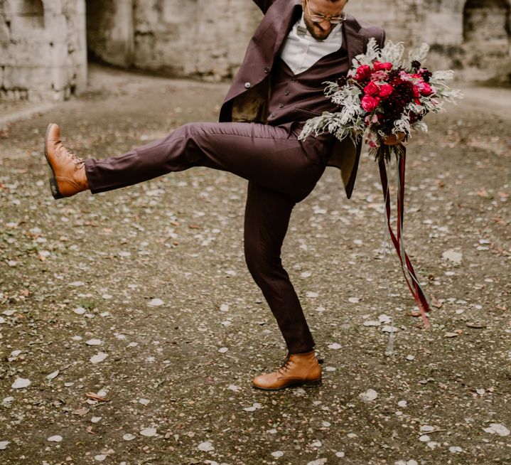 Groom in a burgundy suit holding a red wedding bouquet 