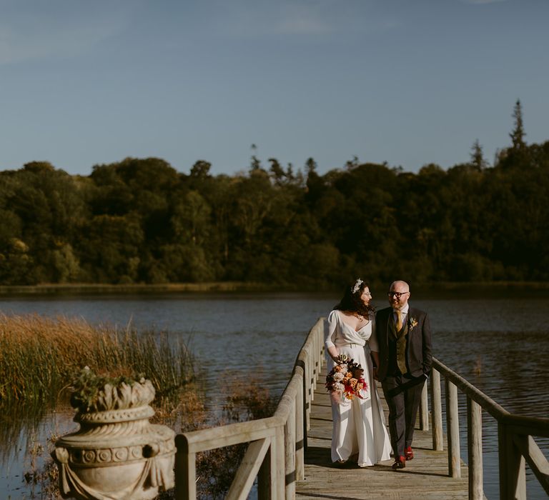 Bride in white Rime Arodaky wedding dress holding pink and white rose and pampas grass bouquet walks along wooden jetty with groom in navy suit and yellow tie