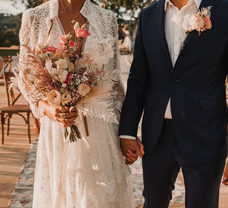 Groom in a navy suit and bride in a bohemian Marylise Bridal gown holding a coral a cream wedding suit