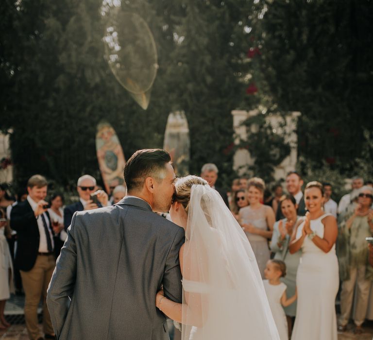 Bride and groom standing in front of their guests, the groom kissing the bride on the head