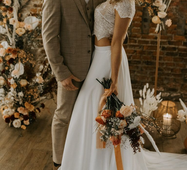 Bride and groom standing nose to nose in front of floral bouquets of roses, the bride is wearing wedding dress separates and carrying a bouquet of mixed roses