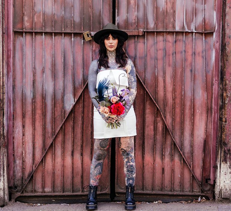 Tattooed bride in a short wedding dress with Dr Marten boots and fedora hat holding a colourful wedding bouquet 