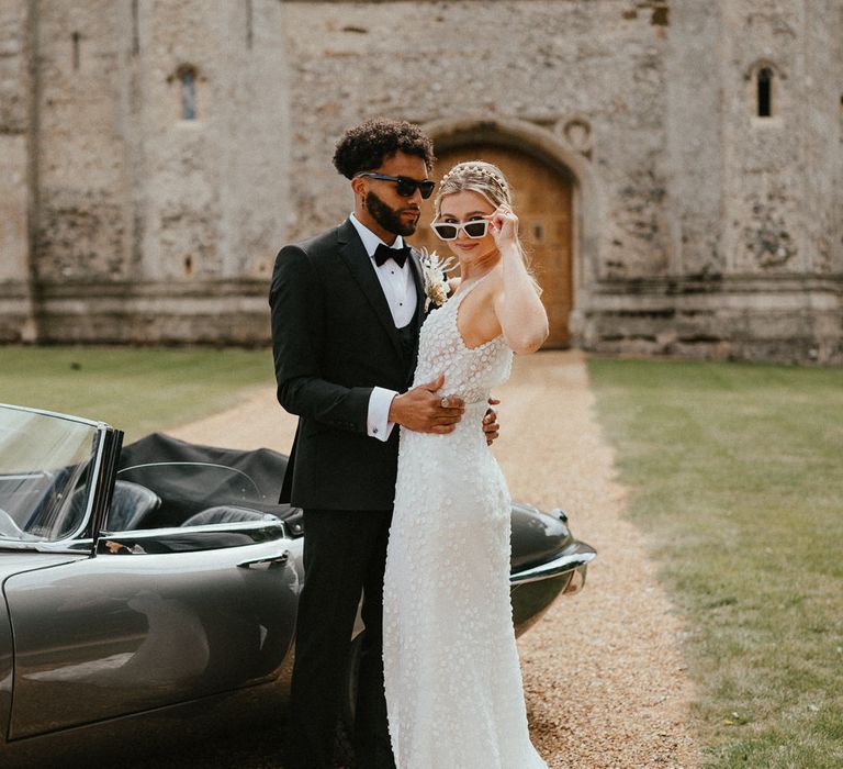 Bride in an appliqué wedding dress, peal headband and white sunglasses standing outside Pentney Abbey with her husband in a tuxedo 