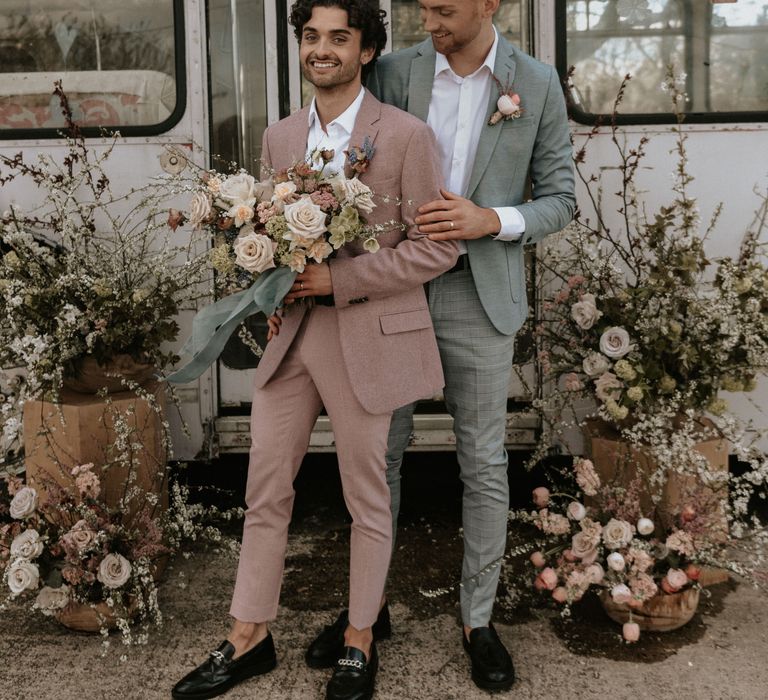 Grooms stand outdoors in front of floral installations whilst smiling and groom holds arm of partner