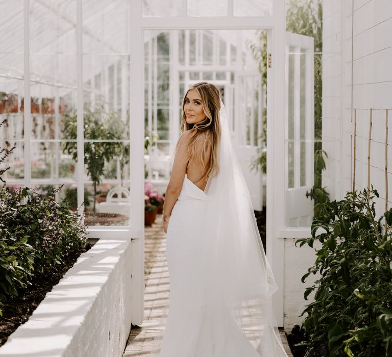 Bride stands within greenhouse whilst looking back toward camera