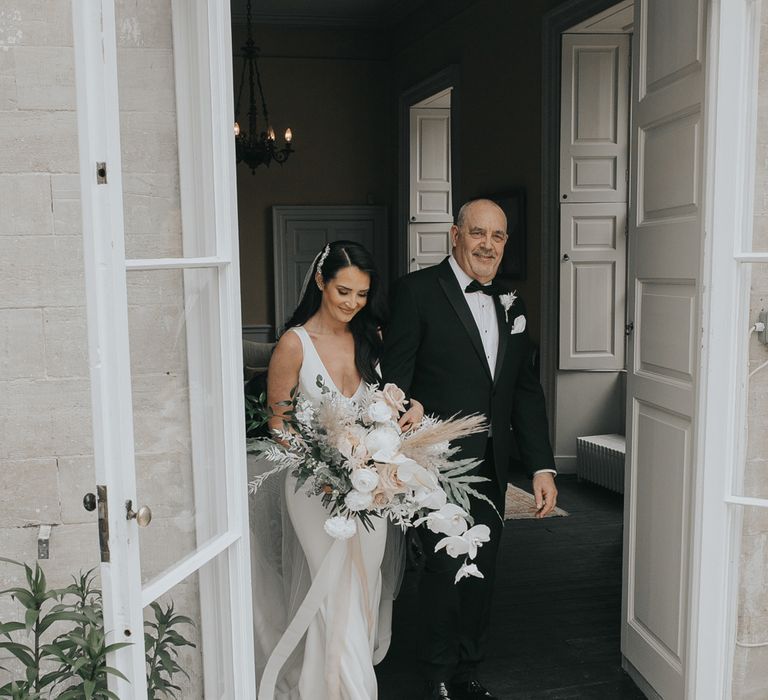 Bride in white Pronovias wedding dress holding large white, pink and green wedding bouquet walks arm in arm with man in tuxedo into the glass conservatory at Came House Dorset for wedding