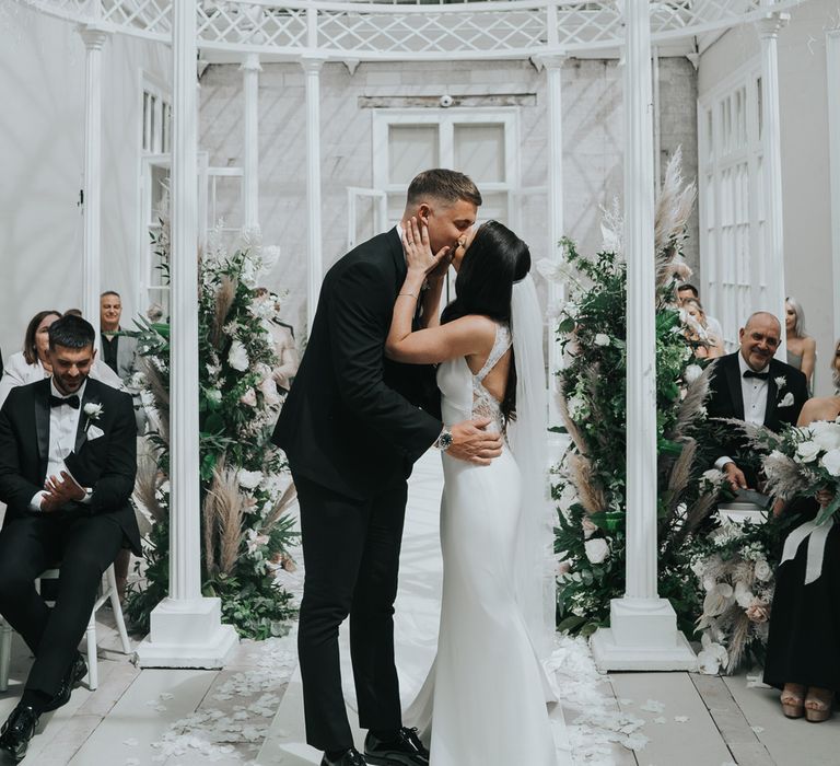 Groom in black tuxedo and bride in white Pronovias wedding dress kiss during wedding ceremony in the glass conservatory at Came House Dorset