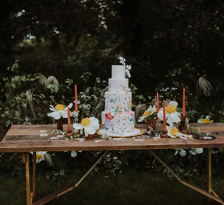 four tier wedding cake with flower illustration resting on a wooden table with wildflower decor 