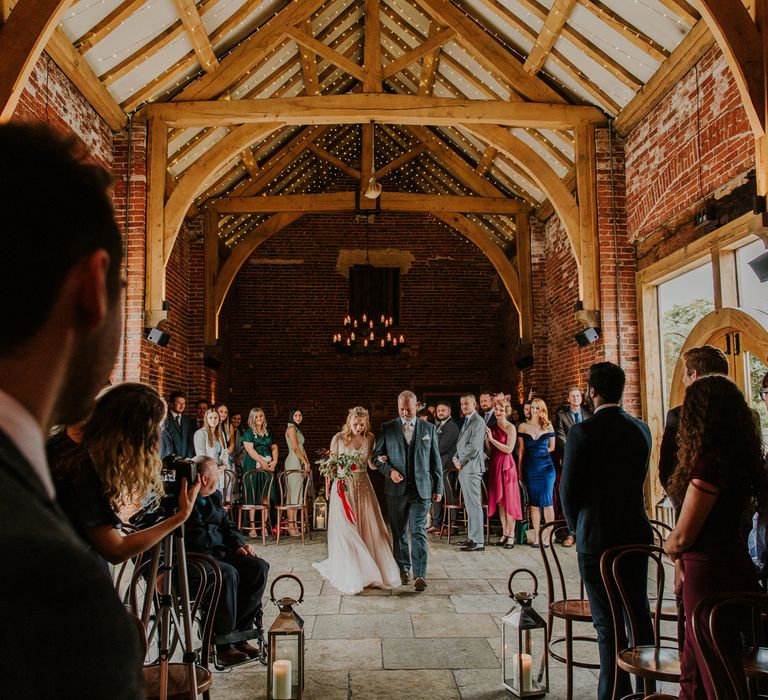 Bride in pearl wedding dress holding red floral bouquet and groom in checked suit hold hands whilst guests watch inside barn after wedding ceremony