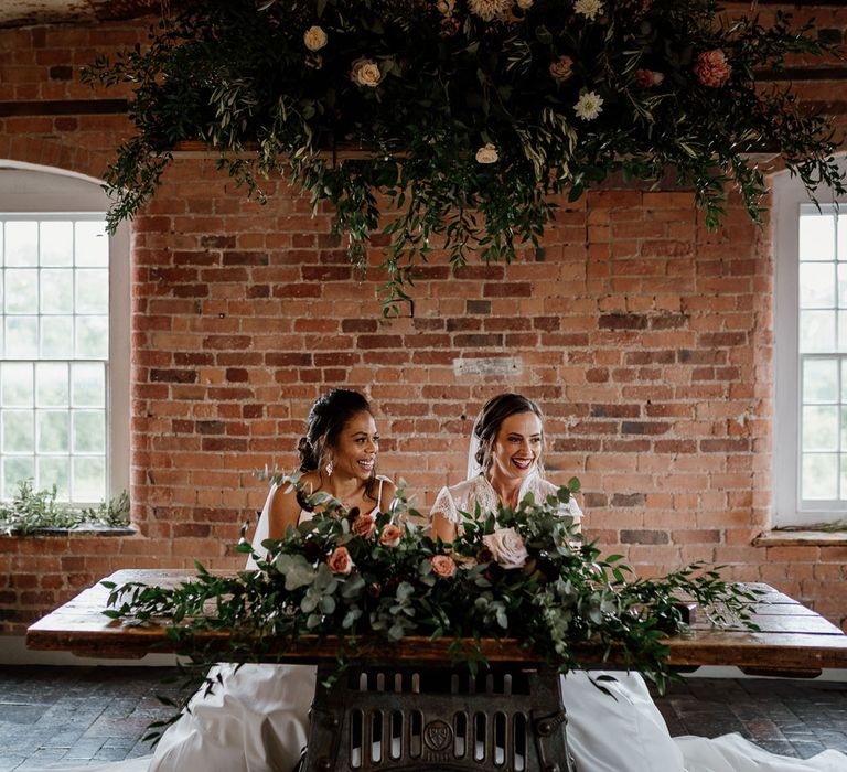 Two smiling brides sit behind wooden table under foliage and flower cloud inside rustic red brick wedding ceremony room at The West Mill Derby