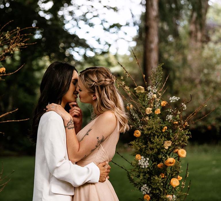 Bride with tattoos in a blush pink wedding dress kissing her bride at the altar in a white trouser suit 