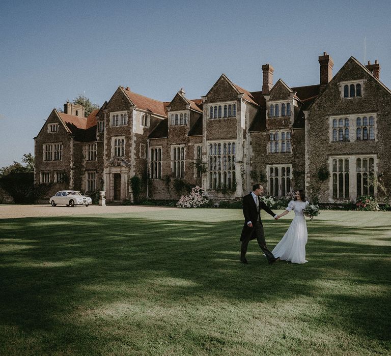 Bride in white lace puffed sleeve Daalarna wedding dress holding white and green bridal bouquet holds hand of with groom in black morning coat and green waistcoat as they walk across the lawns in front of Loseley Park in Surrey