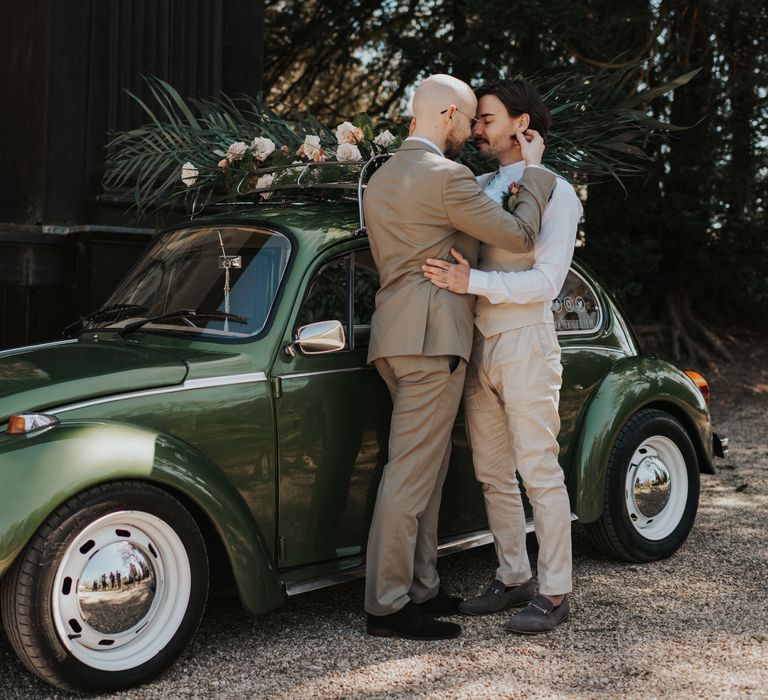 Two grooms in beige and stone suits embracing by a vintage green beetle wedding car with roof rack decorated in foliage