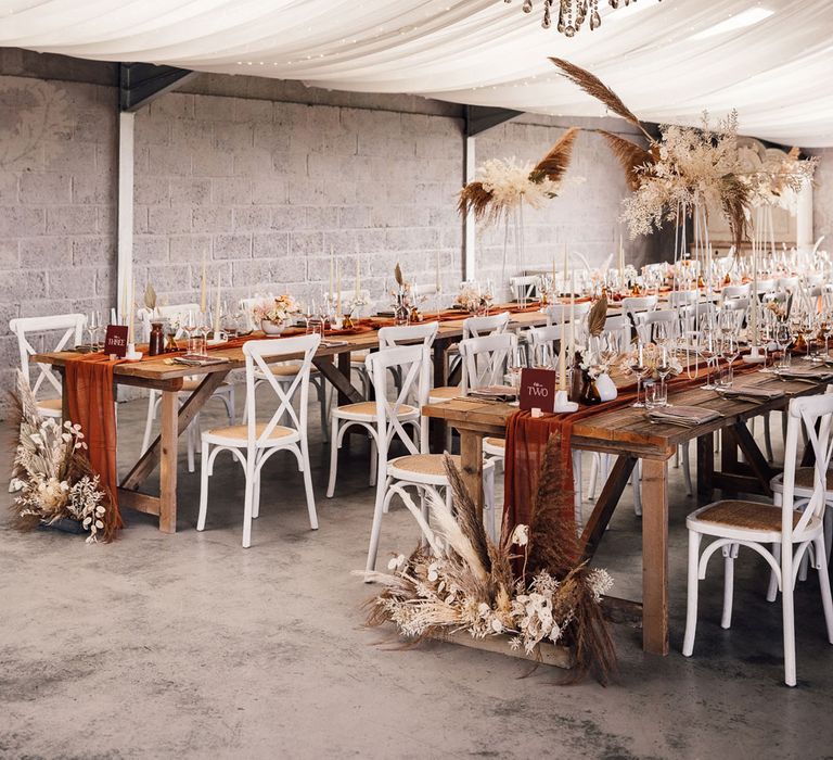 Boho wedding breakfast with long wooden table, burgundy table runner, pampas grass, white chairs and white drapery for summer wedding in Dorset