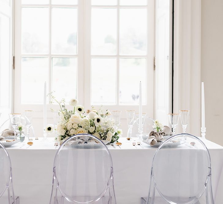 A table with 6 ghost chairs has a white table cloth, white candles and gold rimmed glassware for a black and white wedding reception. 