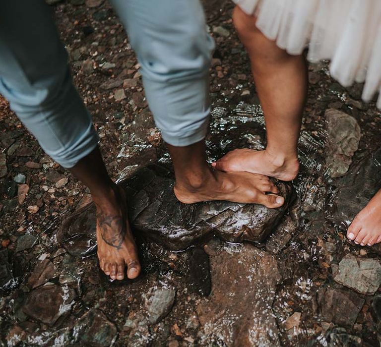 Bride & grooms barefoot feet surrounded by water and pebbles within the woodland