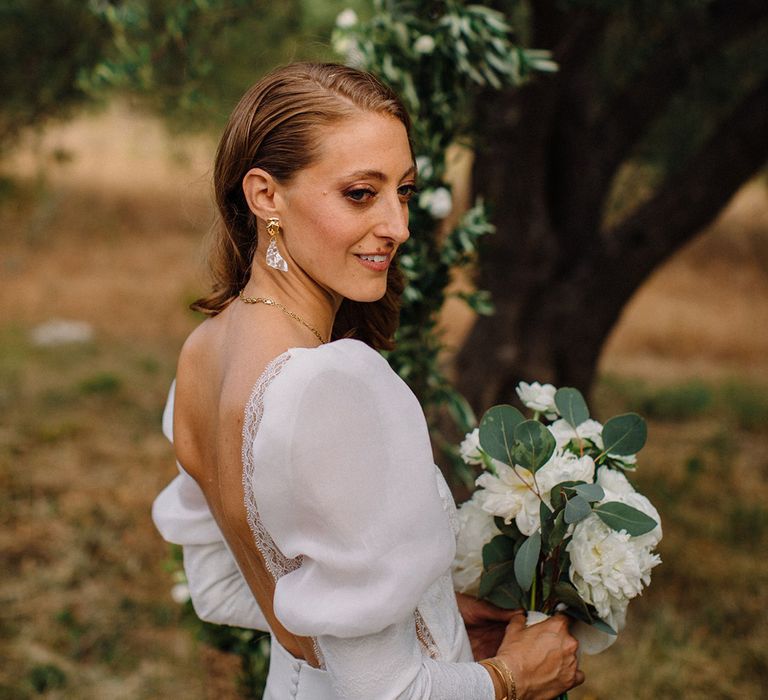 Bride looks to the side as she wears her hair to the side in curls with puff sleeved wedding gown complete with low-cut back