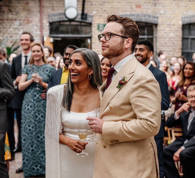 Smiling bride in satin Halfpenny London wedding dress and tasselled bridal cape stands with groom in double breasted linen suit and burgundy tie as they watch speeches outside at Loft Studios London