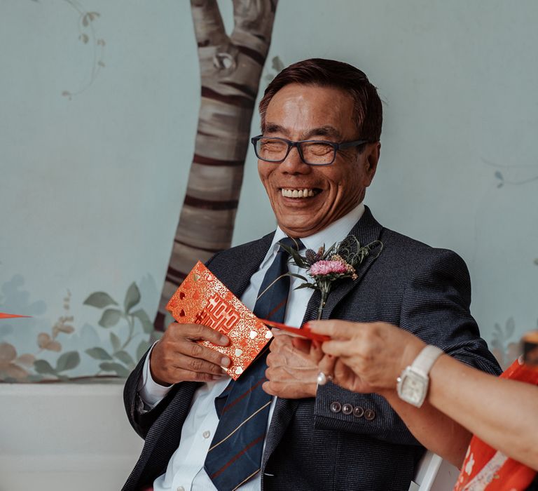 Smiling wedding guest in red trousers and dark blue blazer sits in Victorian summerhouse during traditional Chinese wedding ceremony at Wasing Park wedding