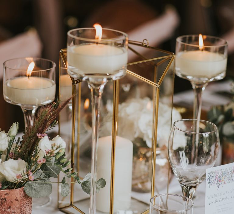 Gold candle box with pillar candle and long stem glass candle holders on white tablecloth next to gold plates and glassware at Tythe Barn wedding with barn wedding flowers