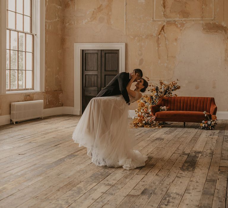 Groom in a tuxedo leaning his bride in a tulle wedding dress over for a kiss with a orange velvet sofa and flower arrangement in the background at Clerkenwell House 