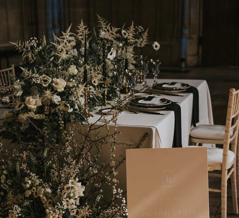 Embossed wedding welcome sign sitting in front of the reception table decorated in white and green flowers 