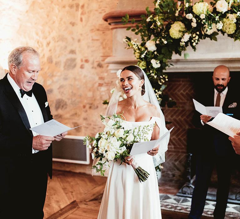 Bride stands at the front of the aisle wearing off the shoulder gown and carrying white floral bouquet