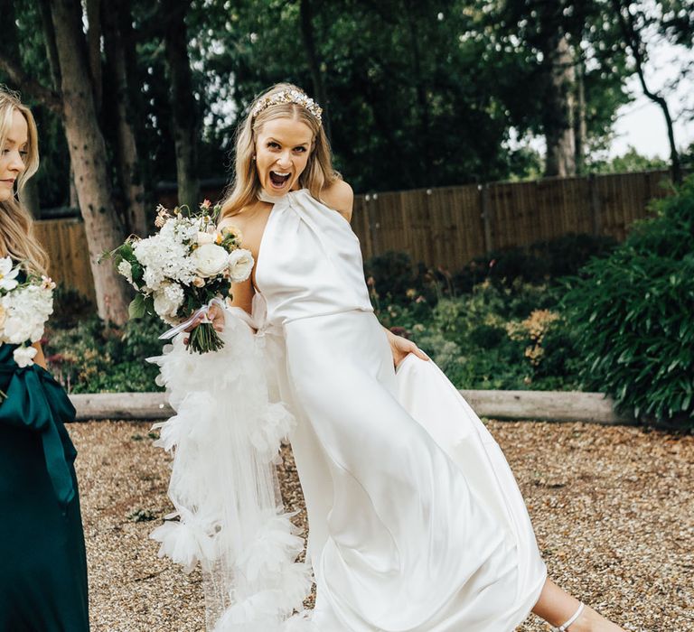 Bride in halterneck Halfpenny London wedding dress and veil with Jimmy Choo wedding shoes kicks out leg as she walks across driveway