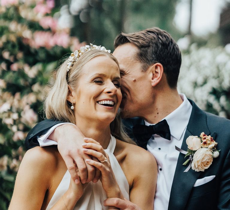 Groom in black tie and floral buttonhole hugs bride in halterneck Halfpenny London wedding dress and bespoke Victoria Percival headband