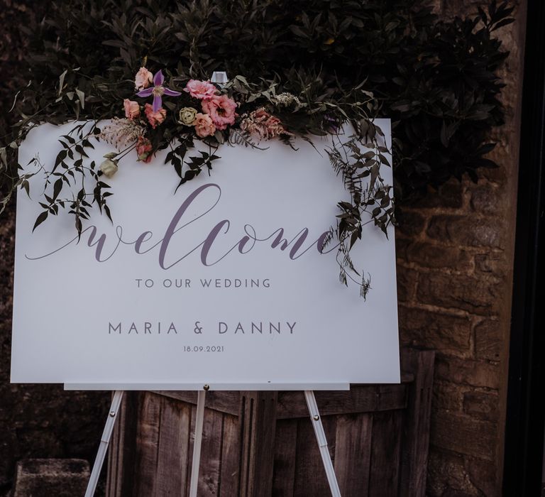 White wedding welcome sign with modern calligraphy topped with floral and foliage garland on white stand 