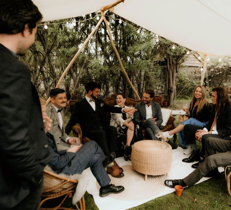 Wedding guests sit under canopy lit with edison light bulb string lights on vintage furniture at rustic wedding