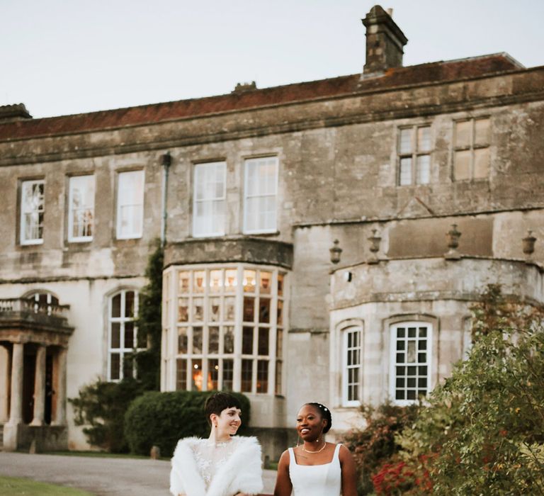 Bride in a faux fur wrap arm in arm with her bride in a minimalist princess wedding dress at their country house wedding 