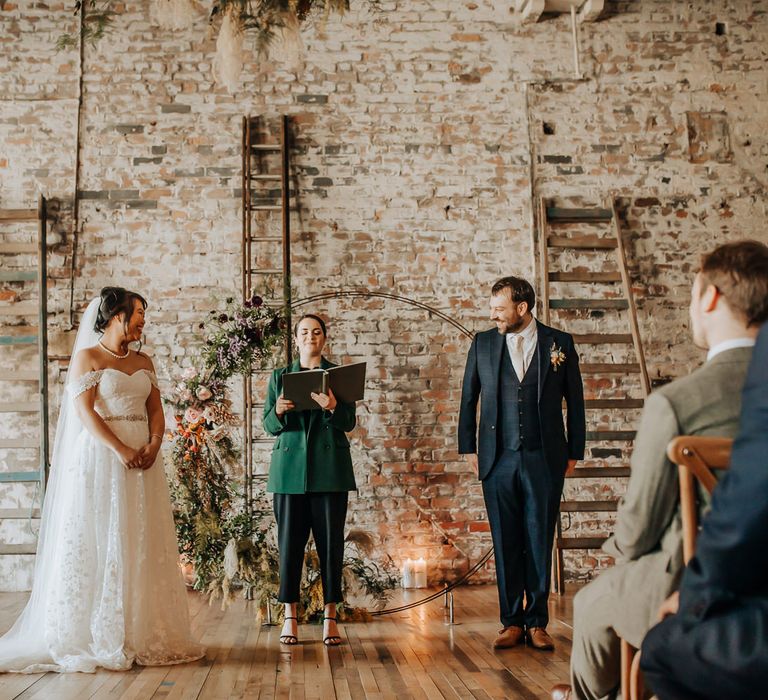 Celebrant in green blazer reads whilst bride in lace bardot wedding dress, pearl necklace and veil stands with groom in blue three piece suit and pink tie during wedding ceremony