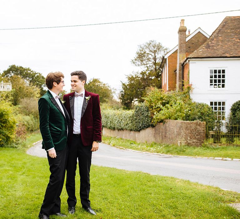 Grooms in velvet tuxedo jackets pose together outside wedding venue in Essex 