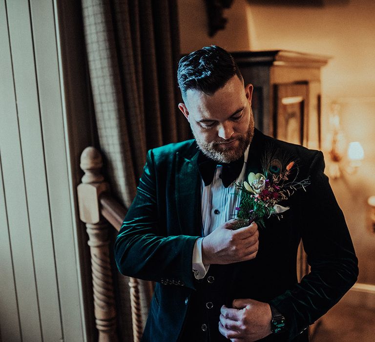 Groom in green velvet suit jacket attaches his large buttonhole with bright flowers and peacock feather 