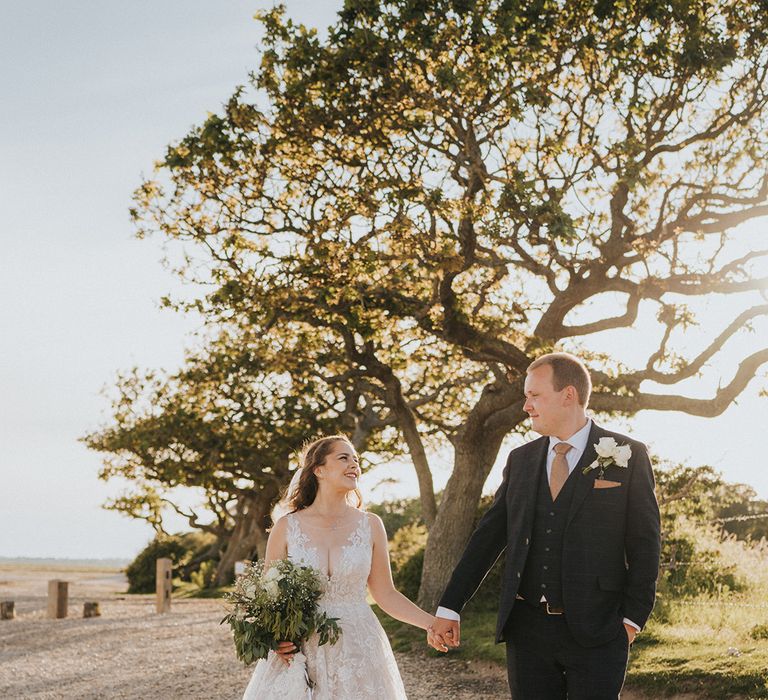 Bride in lace dress and groom in checkered suit hold hands on the private beach at their wedding venue 