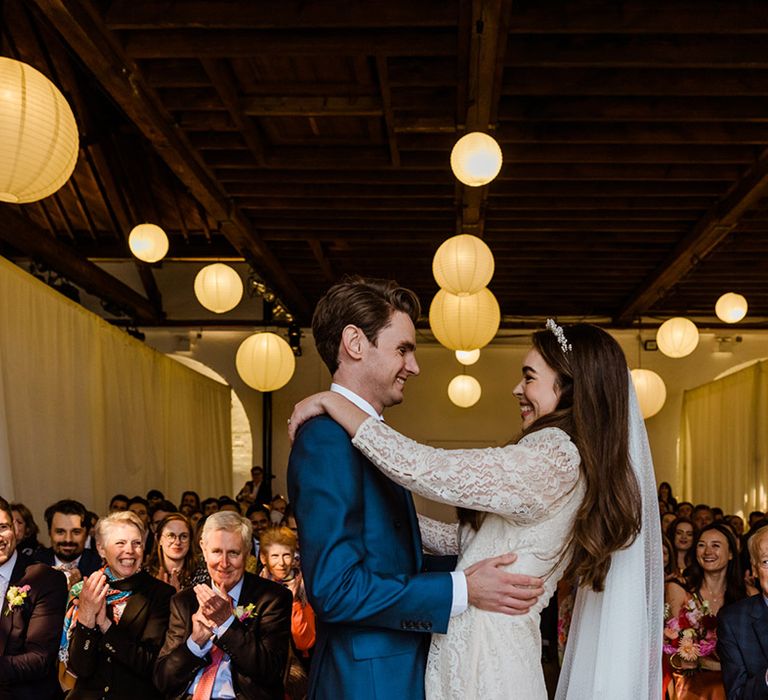 Bride in Rixo dress and pearl headband and groom in blue suit stand at the altar for their wedding with hanging paper lanterns