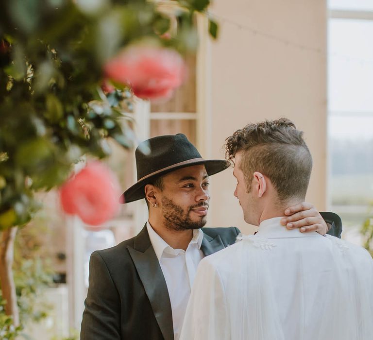 Groom in a black suit and fedora hat embracing his partner in a white groom suit and cape