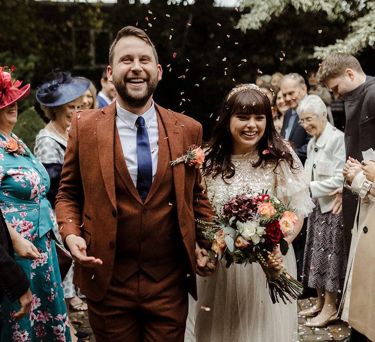 Groom in three piece brown suit and blue tie walks through confetti with the bride in two-piece from Needle and Thread 