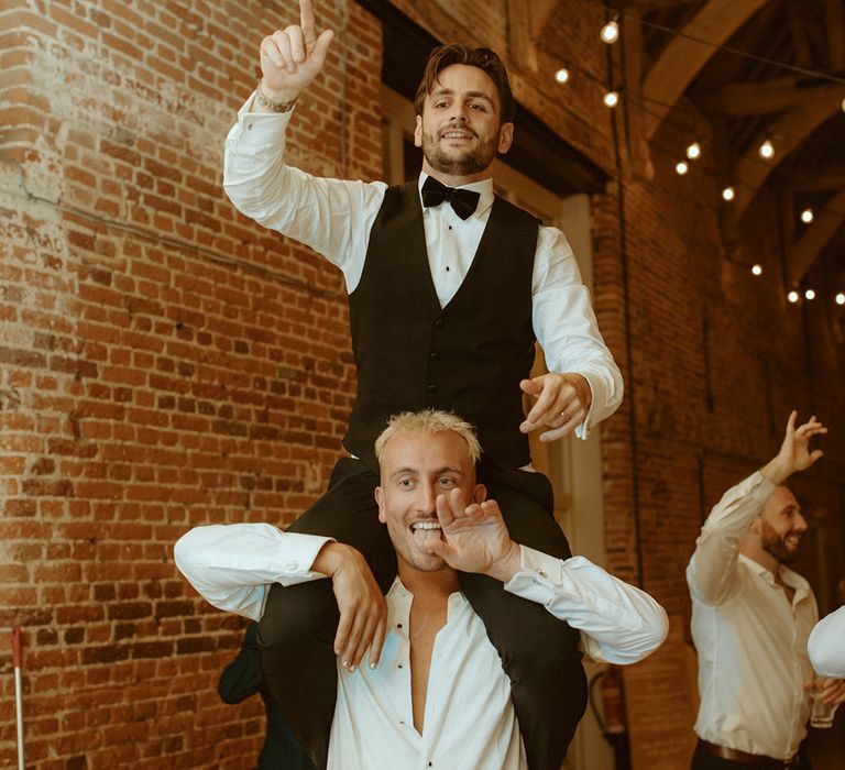 Groom in black waistcoat and bow tie dances on the shoulders of groomsmen in unbuttoned white shirt