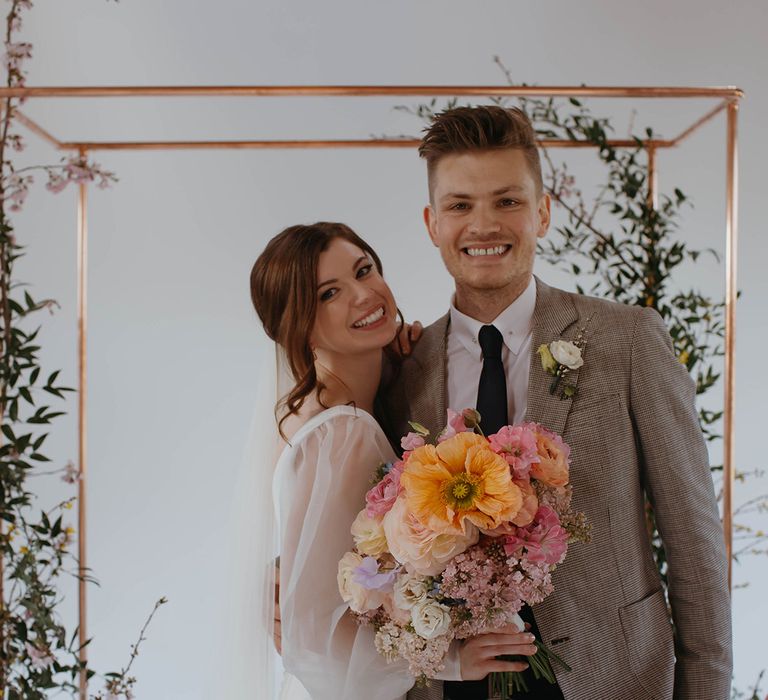 Bride and groom smile together as the bride holds bright spring bouquet with orange giant poppy in front of copper frame decorated with foliage