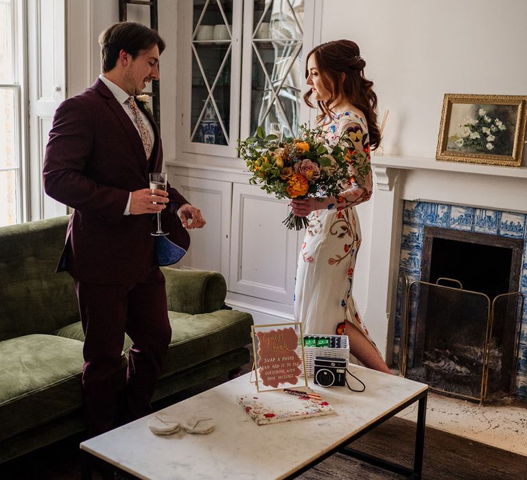 Bride and groom have a glass of champagne together after their ceremony next to table with guest book on 