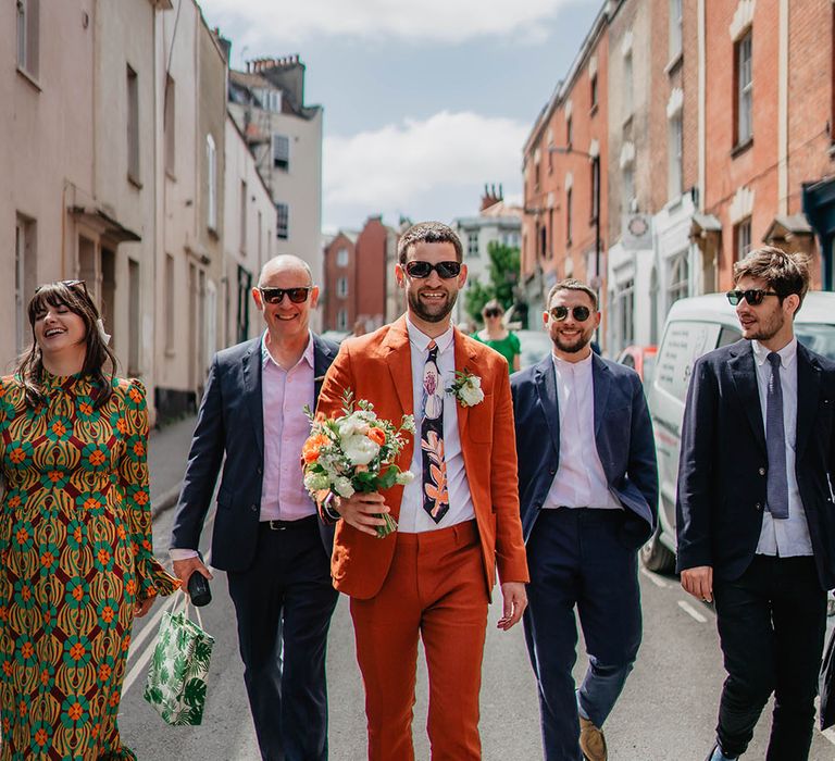 Groom in sunglasses and orange Paul Smith linen suit holds bridal bouquet and walks alongside wedding party in the streets of Bristol 