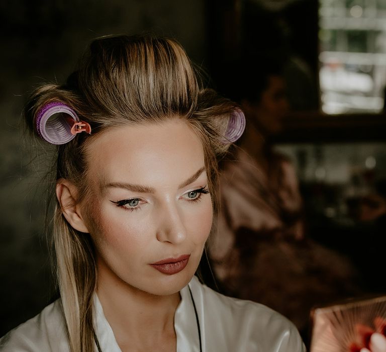 Bride with winged eyeliner holds up a Charlotte Tilbury palette with her hair in rollers as she gets ready